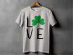 This Sport Grey T-shirt on a hanger is, featuring L♥VE with a shamrock replacing the O, embodies Irish spirit against a plain gray wall for St. Patricks Day.