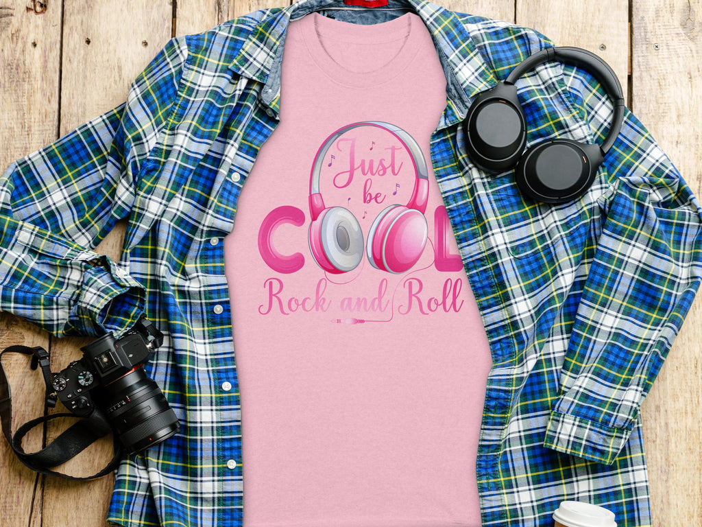 A pink Garment Graphics Rock and Roll T-shirt with Just be Cool, Rock and Roll text and a headphones design lies on a blue and green plaid shirt. Nearby are a black camera, black headphones, and a white paper cup on wood—ideal for music fans.