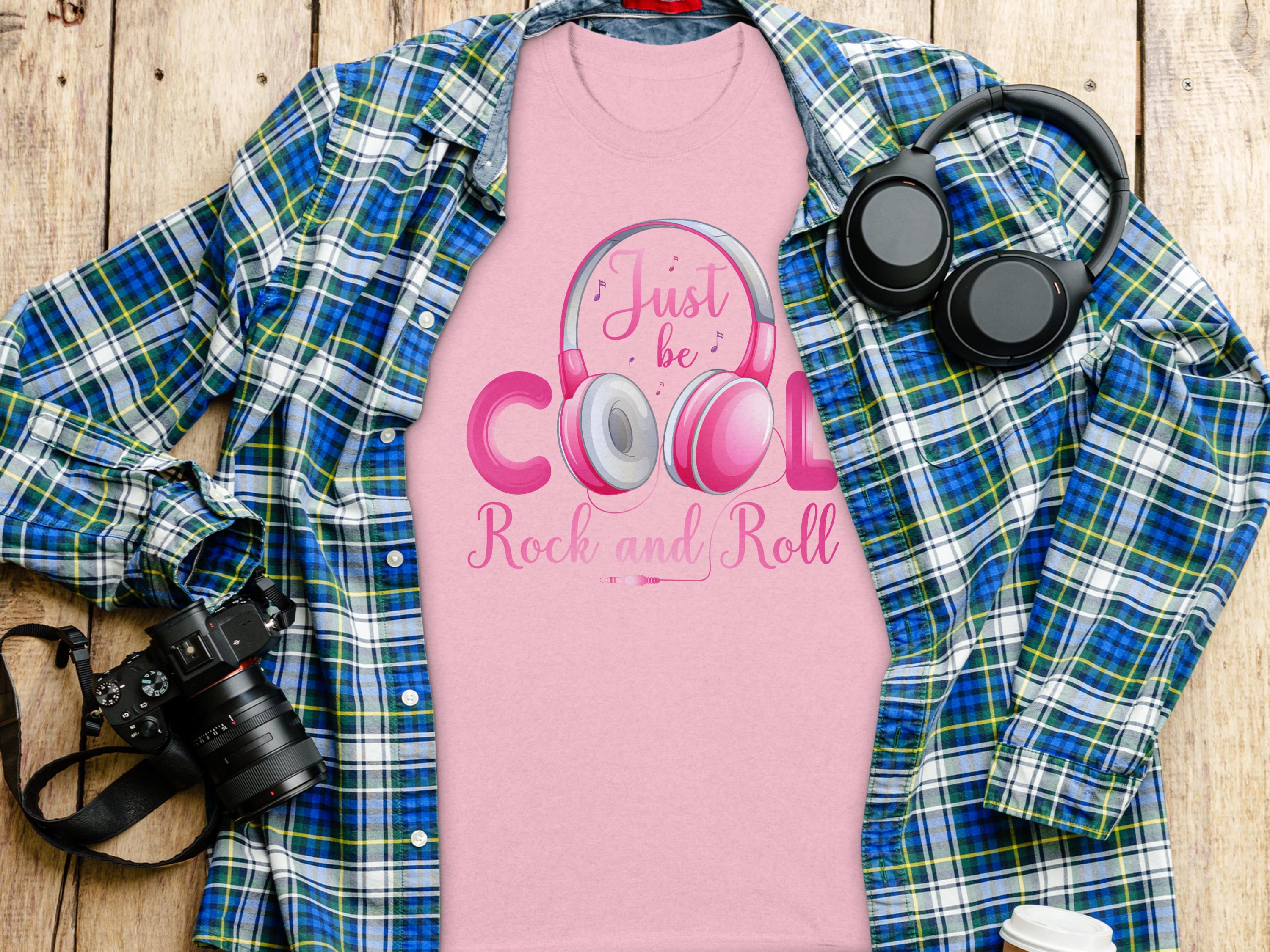 A pink Garment Graphics Rock and Roll T-shirt with Just be Cool, Rock and Roll text and a headphones design lies on a blue and green plaid shirt. Nearby are a black camera, black headphones, and a white paper cup on wood—ideal for music fans.