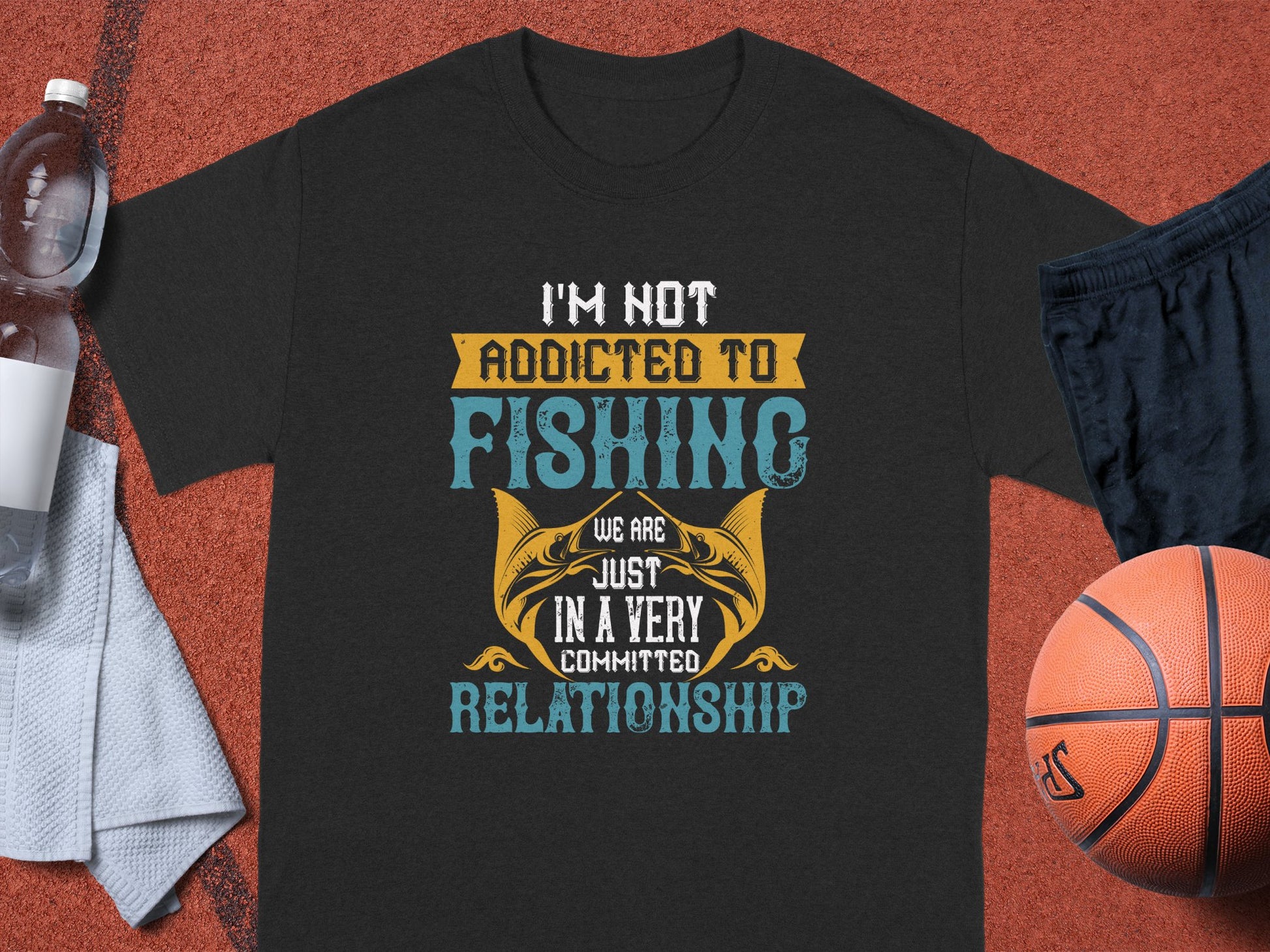 The Garment Graphics fishing T-shirt, set against an orange backdrop, displays the text Im not addicted to fishing, we are just in a very committed relationship alongside fish images. It is surrounded by a water bottle, towel, black shorts, and a basketball—ideal for devoted anglers.