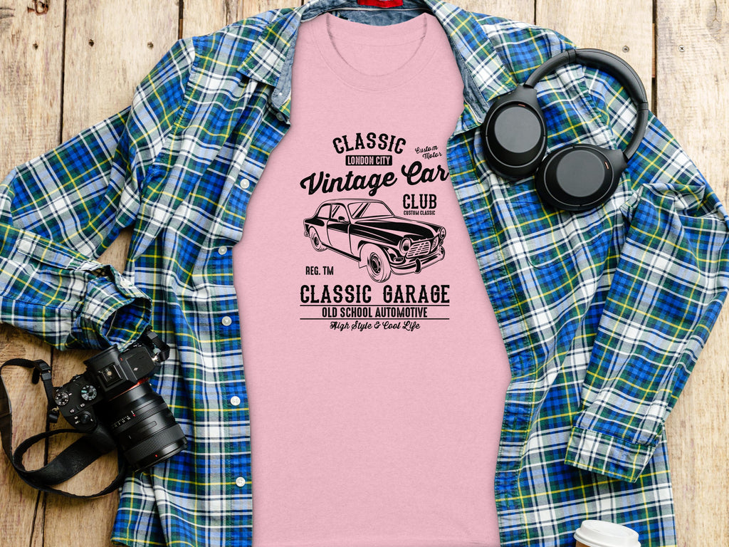 A pink Classic Fit T-shirt from Garment Graphics with a vintage car design and text Classic London City Vintage Car Club and Classic Garage is displayed on a blue plaid shirt, alongside a camera, headphones, and rustic wooden planks in the background.