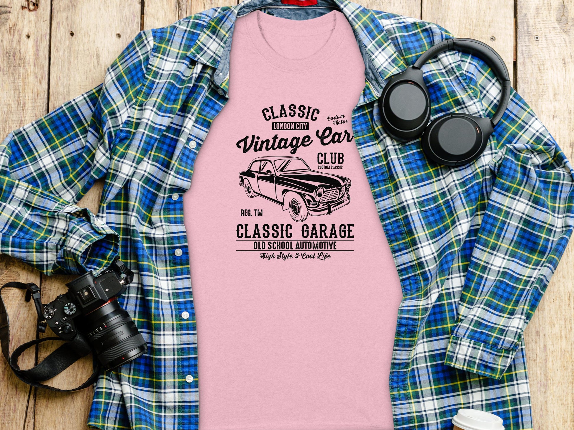 A pink Classic Fit T-shirt from Garment Graphics with a vintage car design and text Classic London City Vintage Car Club and Classic Garage is displayed on a blue plaid shirt, alongside a camera, headphones, and rustic wooden planks in the background.