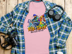 A Garment Graphics pink T-shirt featuring an Artistic Risk Taker Street Dance 10th Final design rests on a plaid flannel shirt, capturing street dance culture. Nearby, a pair of headphones and a camera sit on the wooden surface.