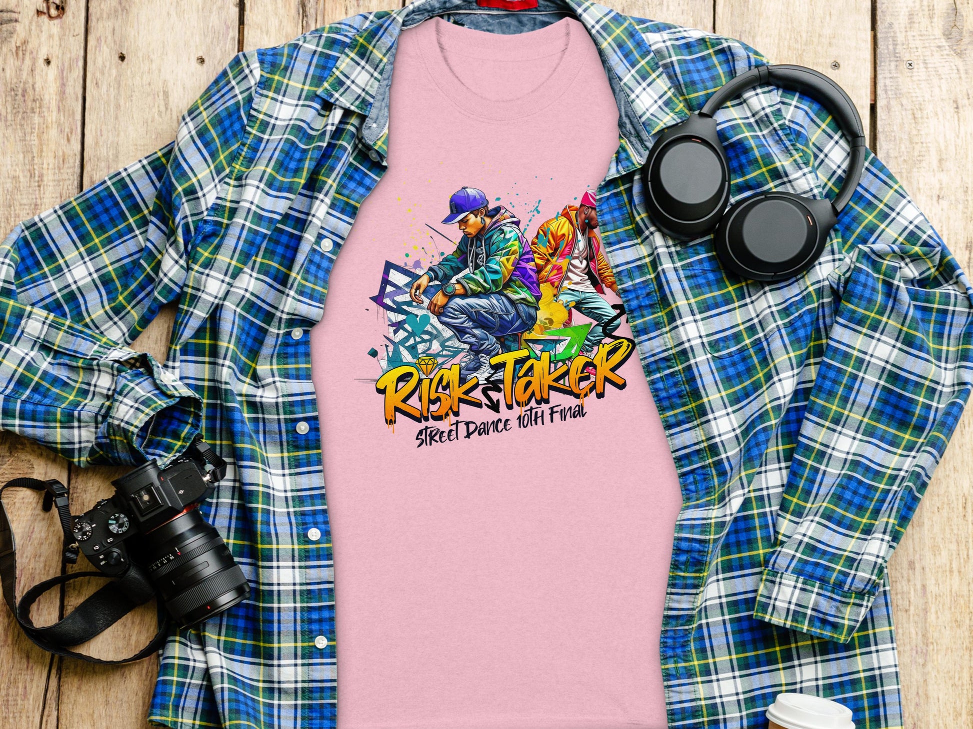 A Garment Graphics pink T-shirt featuring an Artistic Risk Taker Street Dance 10th Final design rests on a plaid flannel shirt, capturing street dance culture. Nearby, a pair of headphones and a camera sit on the wooden surface.