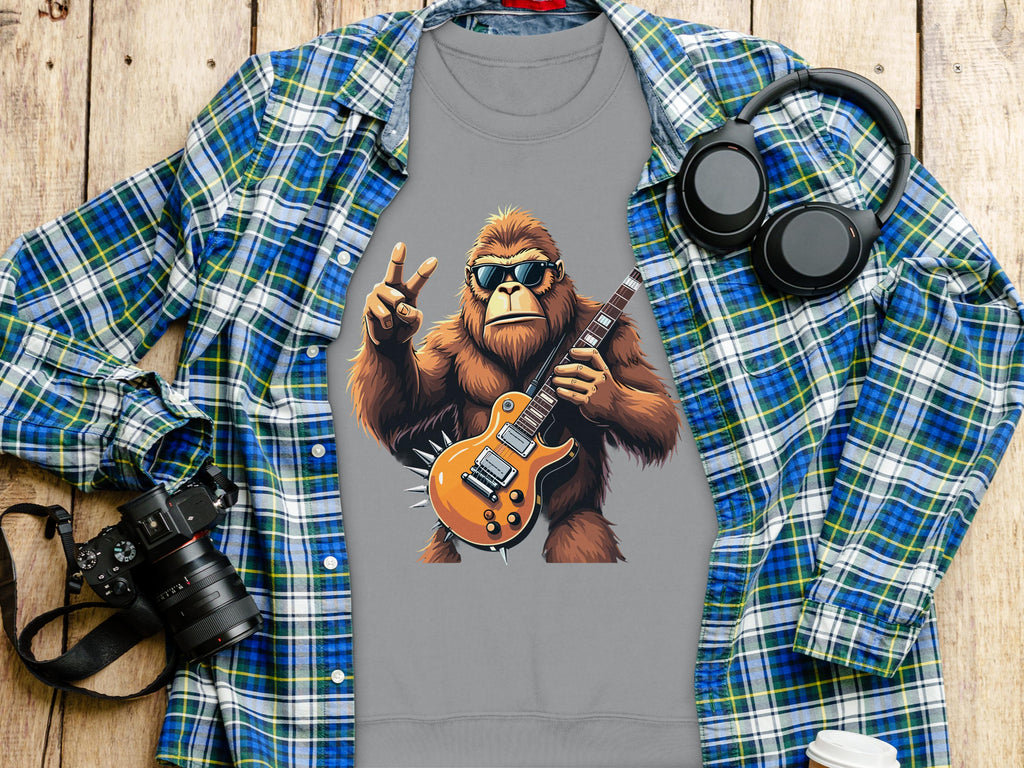 A gray T-shirt from Garment Graphics features a cartoon ape in goggles playing an electric guitar under a plaid shirt, ideal for music lovers. Nearby sit a camera, headphones, and white cup on wood.