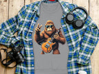 A gray T-shirt from Garment Graphics features a cartoon ape in goggles playing an electric guitar under a plaid shirt, ideal for music lovers. Nearby sit a camera, headphones, and white cup on wood.