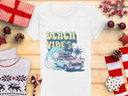 Garment Graphics presents a retro t-shirt showcasing a surf design with a vintage car, surfboard, and palm tree. The Beach Vibes text adds charm to the wooden backdrop adorned with holiday decorations like wrapped gifts, pine cones, and candy canes.