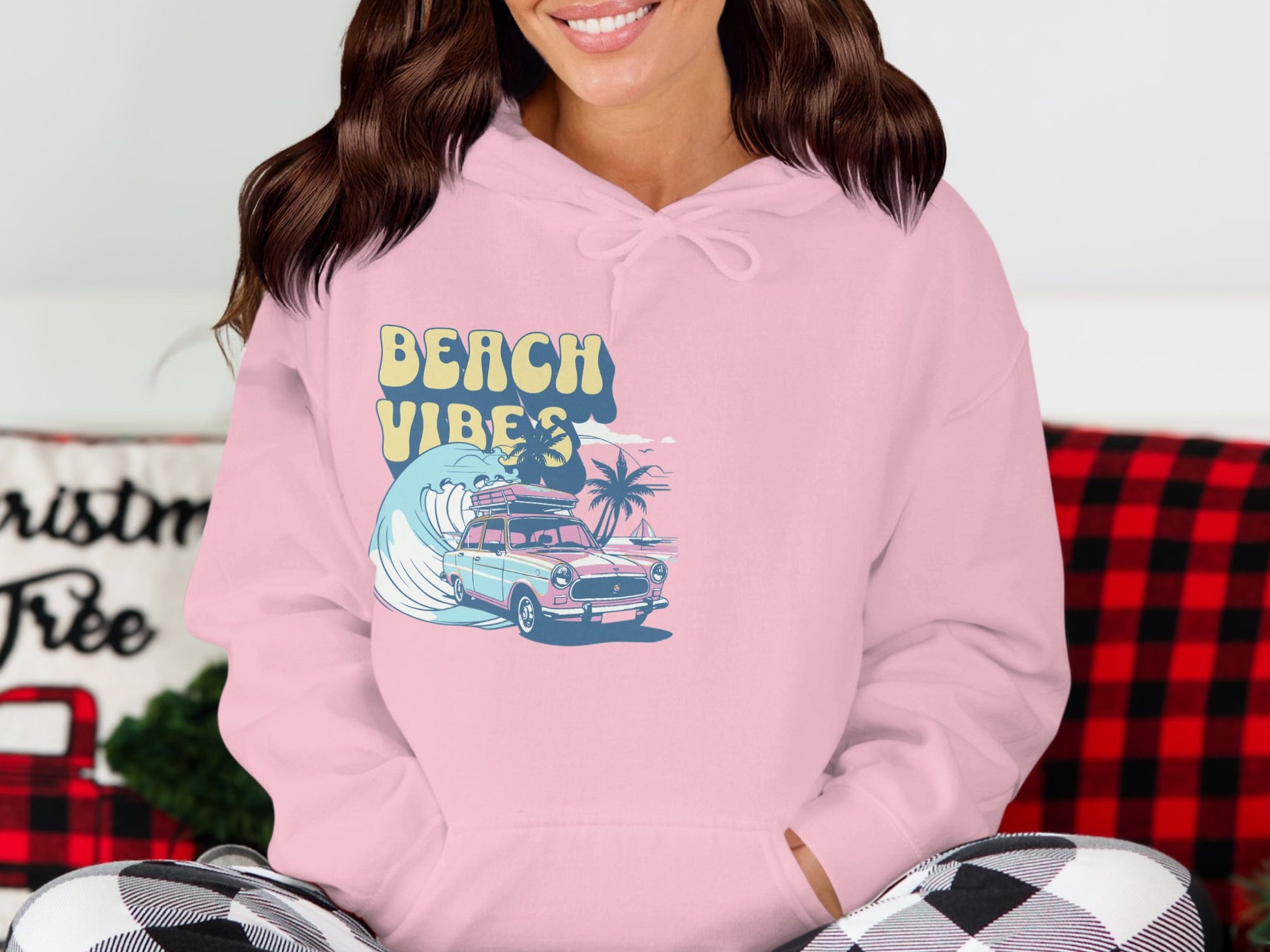 A person wears a retro beach style pink hoodie from Garment Graphics, featuring BEACH VIBES above a car, palm trees, and waves graphic design. Seated in a room with a checked blanket in the background, the vibrant hoodie stands out.