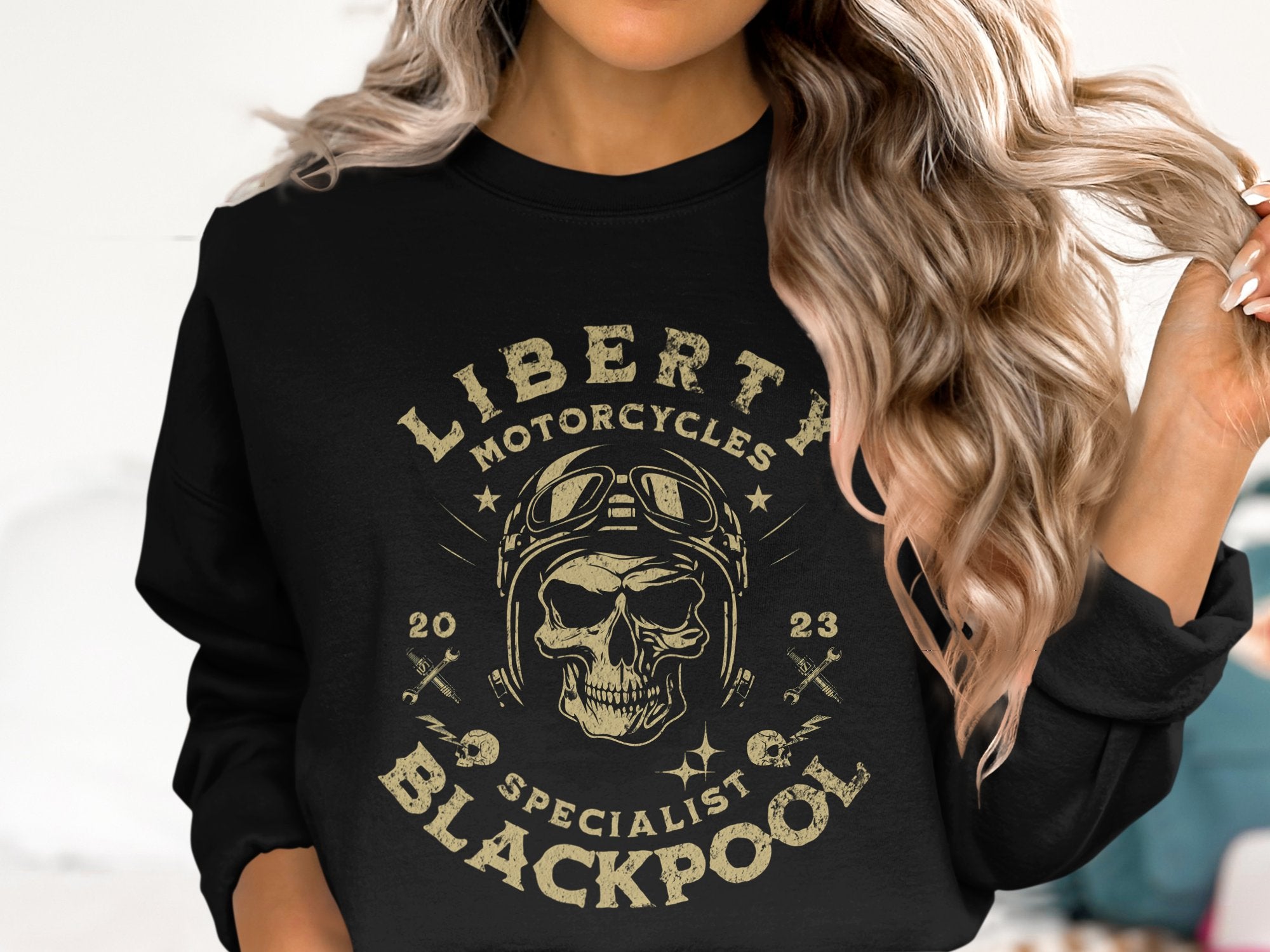 A person with wavy blonde hair is rocking a biker look in a black Garment Graphics sweatshirt featuring a skull with goggles and the text Liberty Motorcycles Specialist Blackpool 2023, while holding a strand of hair.