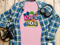 Garment Graphics presents a pink t-shirt with Loud & Proud, Lets Make Some Noise! graphic, displayed on wood next to a blue and green plaid shirt. Ideal for football t-shirt fans, it pairs perfectly with black headphones and a camera, highlighting your passion.