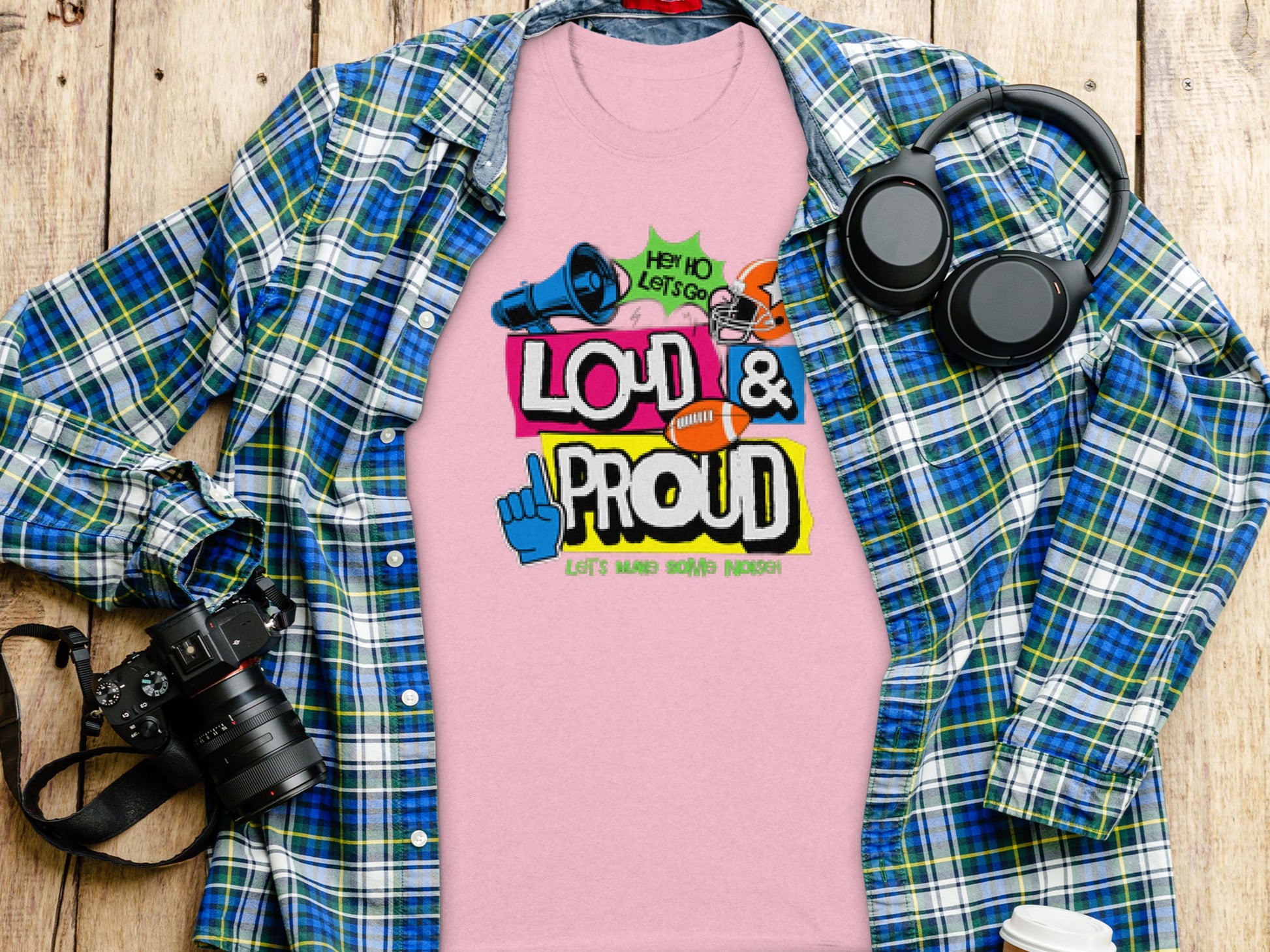 Garment Graphics presents a pink t-shirt with Loud & Proud, Lets Make Some Noise! graphic, displayed on wood next to a blue and green plaid shirt. Ideal for football t-shirt fans, it pairs perfectly with black headphones and a camera, highlighting your passion.