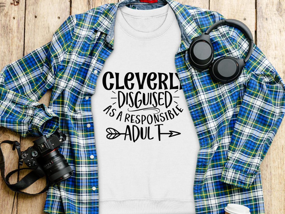 A Garment Graphics white shirt with Cleverly disguised as a responsible adult lies on wood, surrounded by a plaid shirt in blue and green, a camera, headphones, and a coffee cup. With unisex sizing, it’s the perfect humor wardrobe addition.