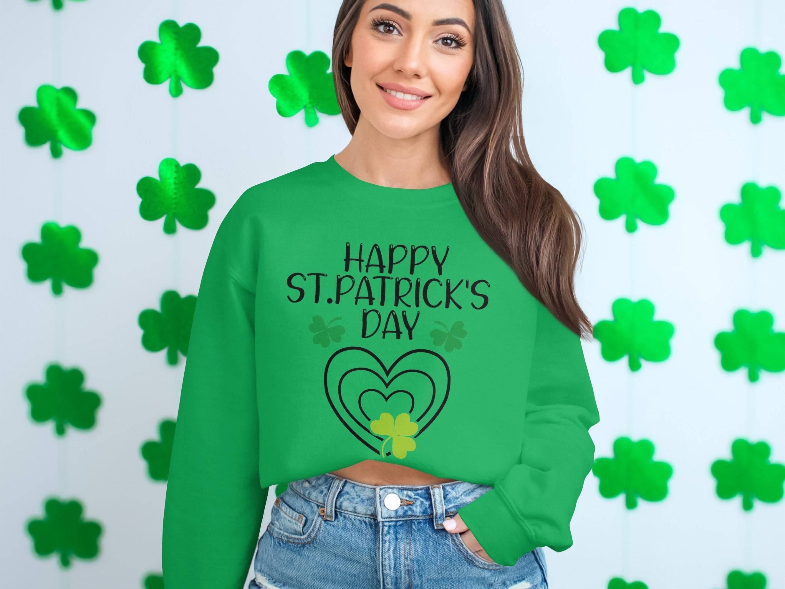 Wearing a Garment Graphics festive sweatshirt with Happy St. Patricks Day and a heart design, a person stands by a wall of green shamrocks. Their long brown hair flows over denim jeans, perfectly embodying the spirit of the day in clover-themed attire.