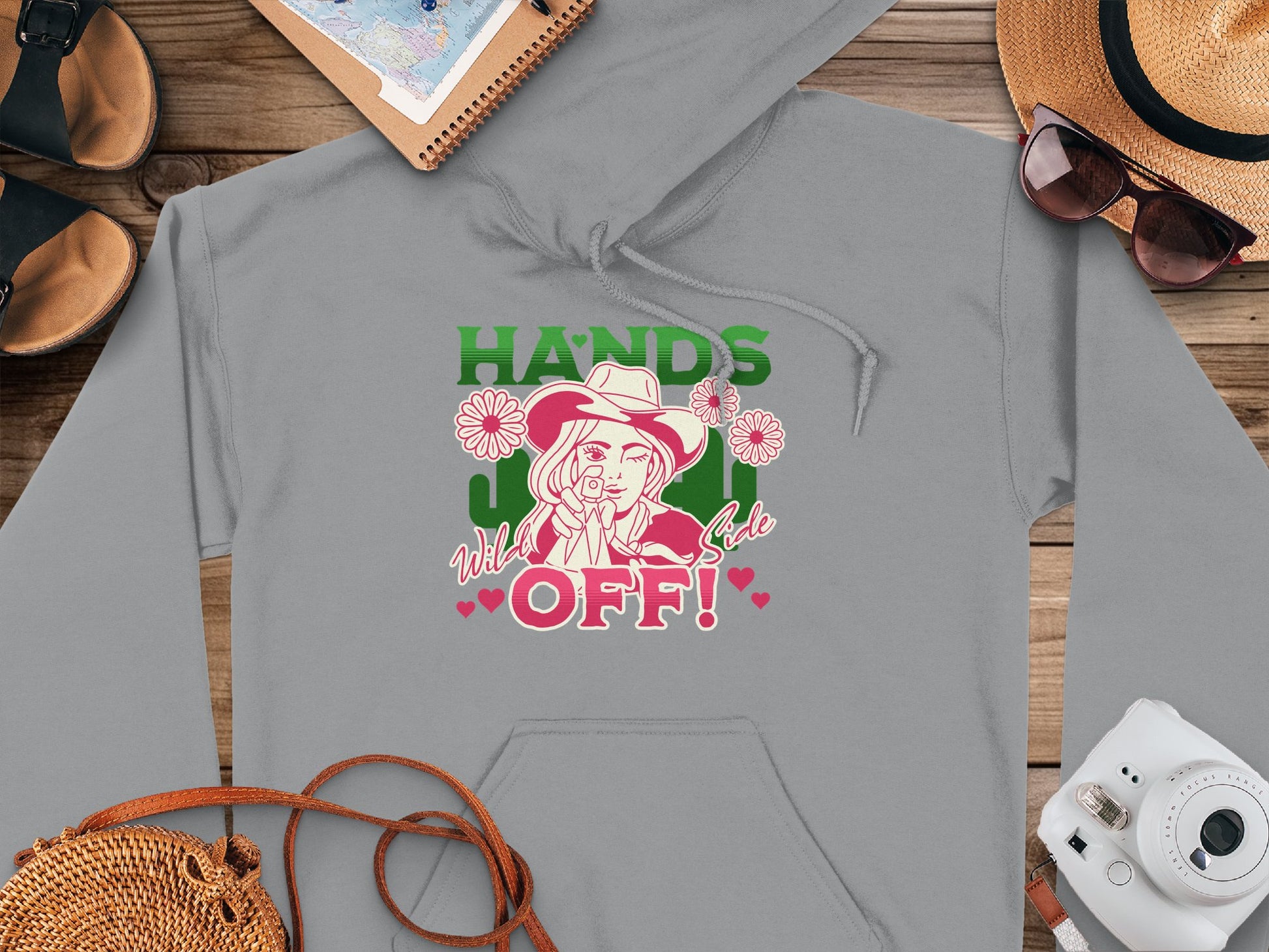 Garment Graphics presents a gray hoodie with a bold statement design of a hat-wearing figure pointing amid flowers. HANDS OFF! is in green and pink, with subtle wild and cute text. Sandals, hat, sunglasses, notebook, and camera nearby make it perfect for Graphic Hoodie enthusiasts seeking flair.