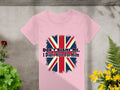 The Garment Graphics Dont Blame Me I Didnt Vote For Him T-shirt boasts a bold distressed Union Jack design on pink, 100% cotton fabric, set against a concrete backdrop with vibrant green and yellow plants for a striking statement.