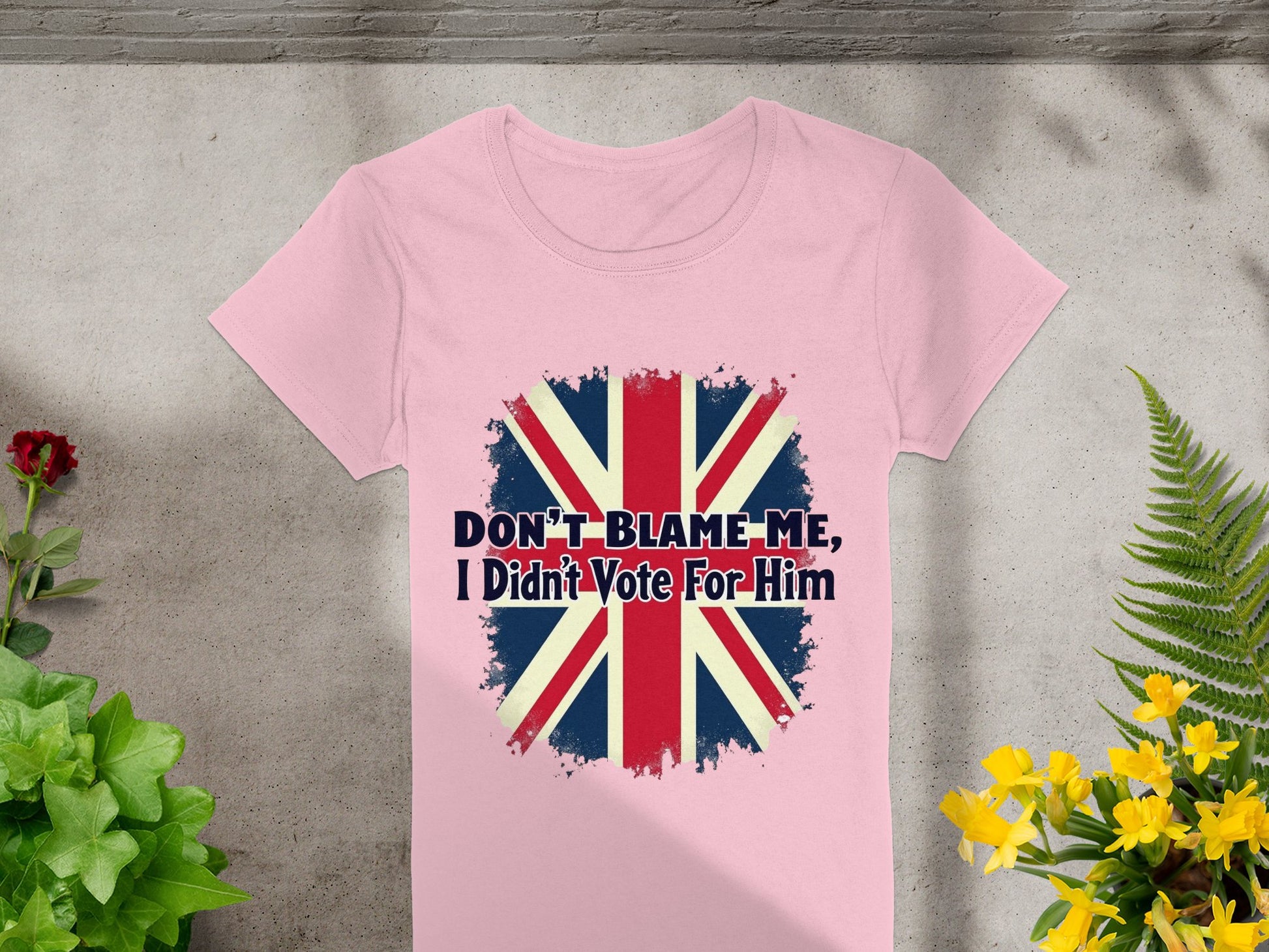 The Garment Graphics Dont Blame Me I Didnt Vote For Him T-shirt boasts a bold distressed Union Jack design on pink, 100% cotton fabric, set against a concrete backdrop with vibrant green and yellow plants for a striking statement.
