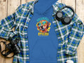 Garment Graphics blue hoodie displays a dog in sunglasses with the text Pawsitive Stylish over a plaid shirt on wood. Nearby are headphones, a camera, a smartphone, and a takeaway coffee cup.