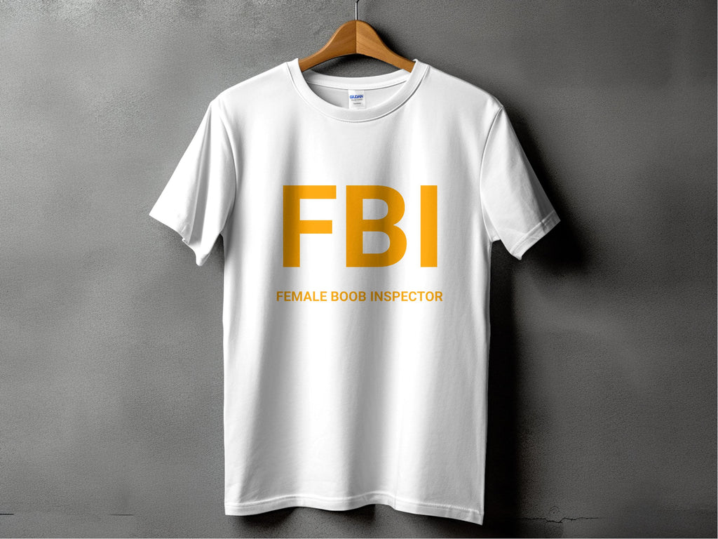 A white T-shirt from Garment Graphics humorously hangs on a wooden hanger against a gray background, flaunting bold yellow FBI text with the playful caption Female Boob Inspector below—ideal for those with a cheeky sense of style.
