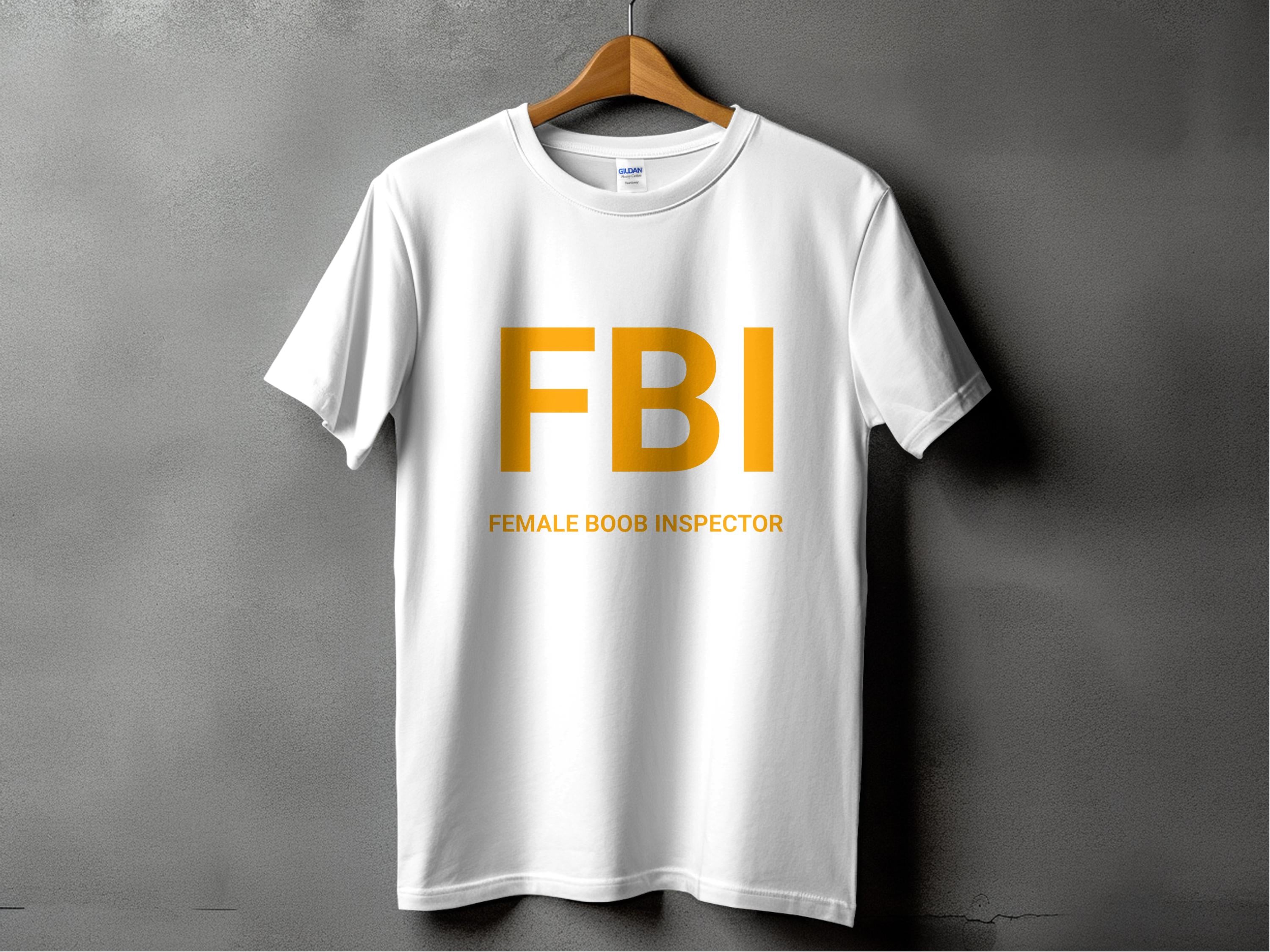 A white T-shirt from Garment Graphics humorously hangs on a wooden hanger against a gray background, flaunting bold yellow FBI text with the playful caption Female Boob Inspector below—ideal for those with a cheeky sense of style.