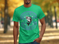 Wearing a green Garment Graphics tee adorned with a fish design and Fishing Hair Dont Care text, a man stands before autumn-colored trees that frame a blurred park path, enhancing the tees unique charm.