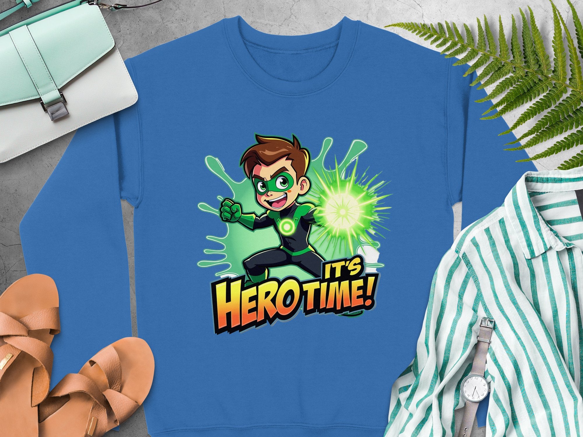 The Garment Graphics sweatshirt features a cartoon superhero child with glowing powers and the bold text Its Hero Time! in unisex sizing, displayed alongside sandals, a striped shirt, watch, purse, and ferns on a gray background.