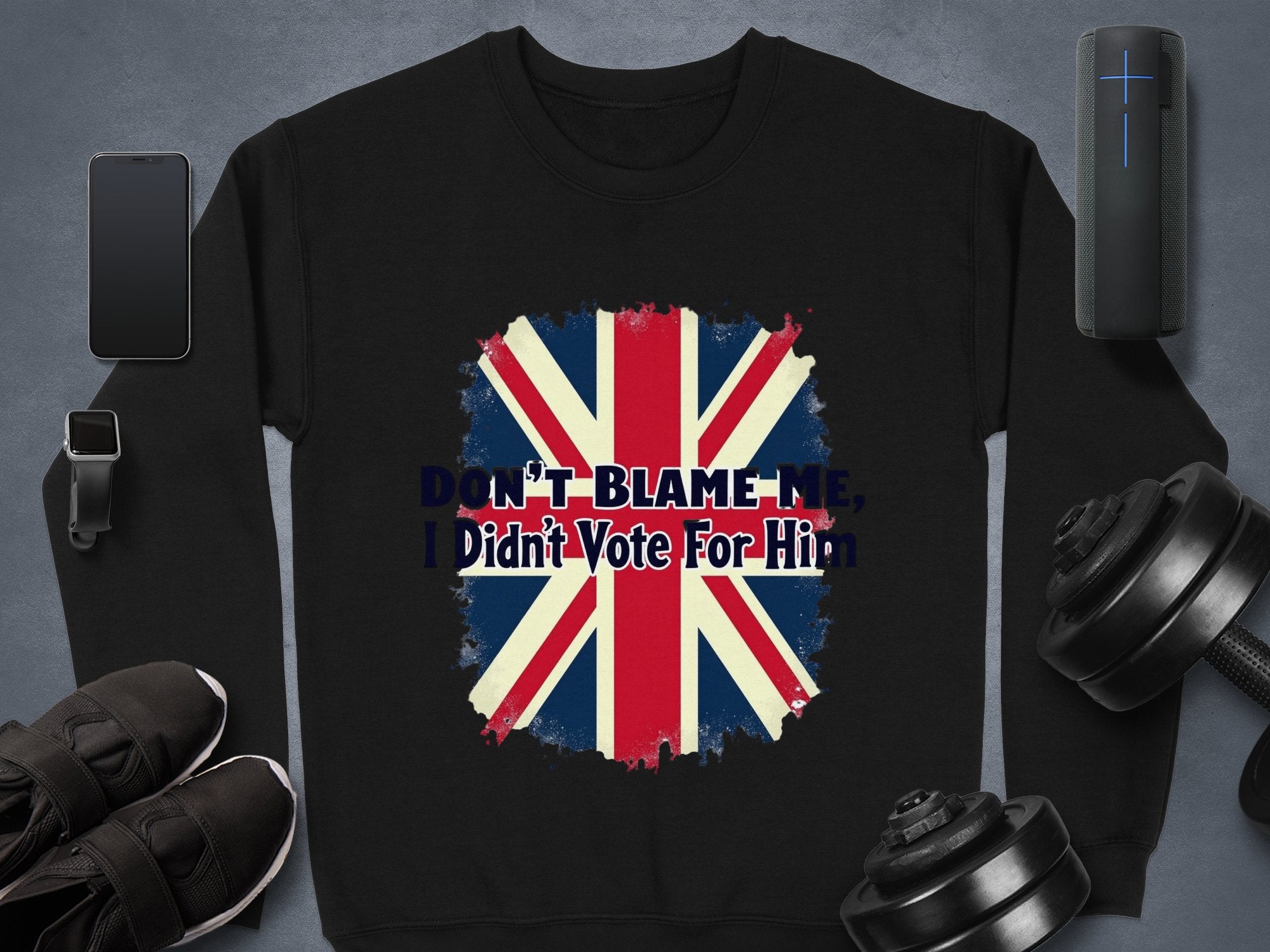 Garment Graphics black sweatshirt showcases a bold, distressed Union Jack and the phrase DONT BLAME ME, I Didnt Vote For Him, set on a gray surface amidst a smartphone, smartwatch, wireless speaker, black sneakers, and dumbbells.