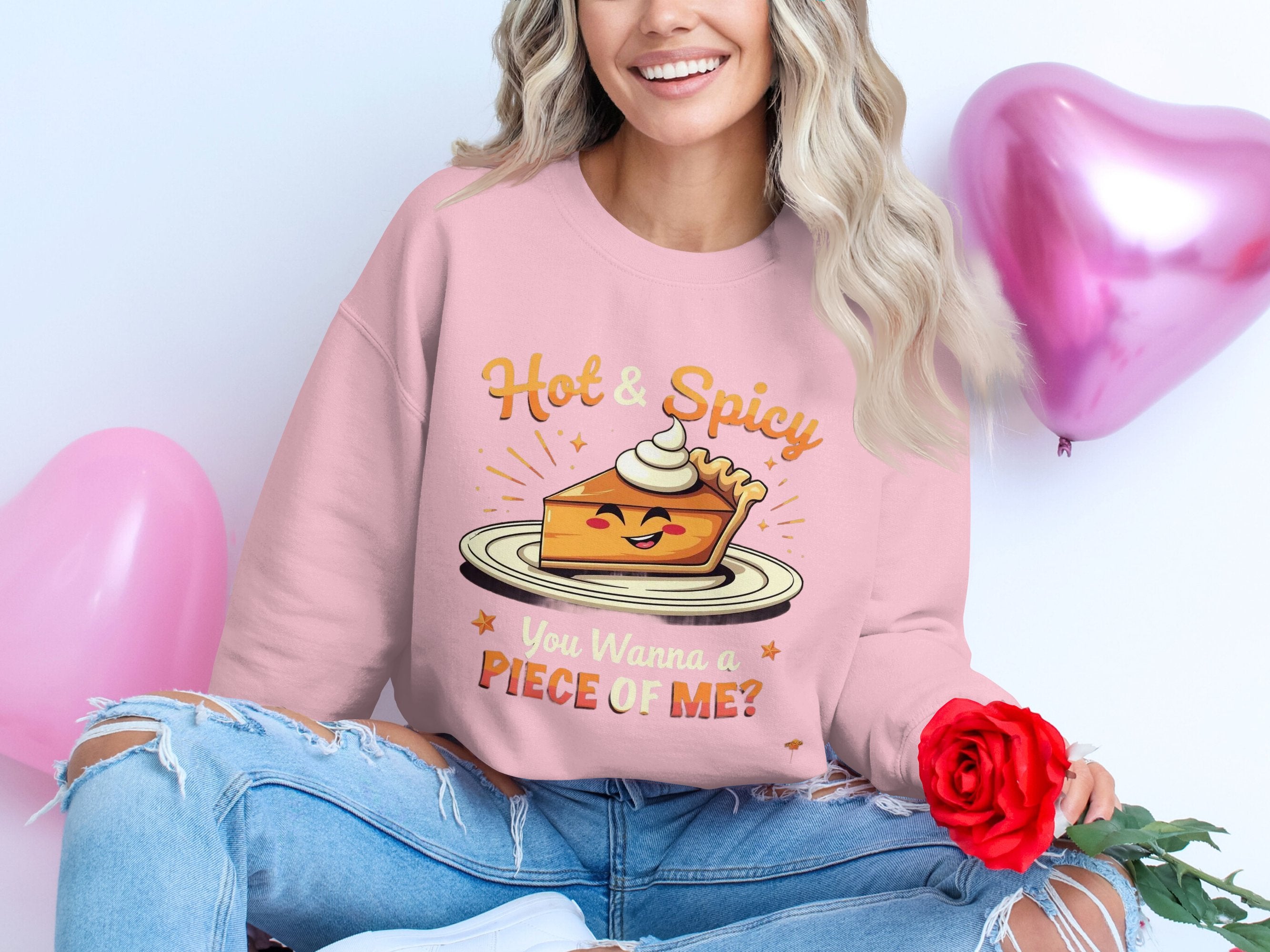 Clad in a Garment Graphics unisex sweatshirt featuring Hot & Spicy, You Wanna a Piece of Me? pie graphic, and ripped jeans, a person holding a red rose smiles radiantly while seated before pink heart balloons.
