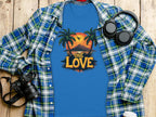 The Garment Graphics blue T-shirt, featuring a One Love design with palm trees and a tropical sunset, is perfect for beach lovers. Its displayed on a wooden surface with a plaid shirt, headphones, a camera, and a white cup with a lid nearby.