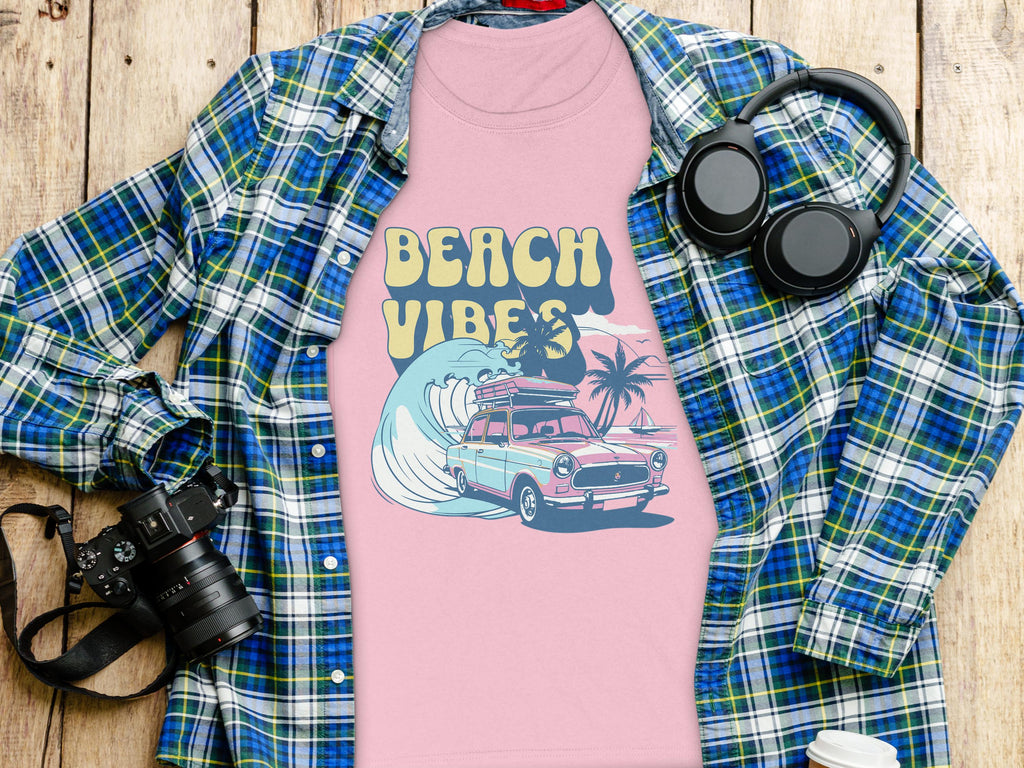 A Garment Graphics retro pink T-shirt with Beach Vibes in vibrant yellow showcases a surf design with a car, surfboard, and palm trees by crashing waves. Its placed on a blue plaid shirt surrounded by headphones, a camera, and a white cup on a wooden surface.