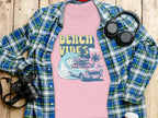 A Garment Graphics retro pink T-shirt with Beach Vibes in vibrant yellow showcases a surf design with a car, surfboard, and palm trees by crashing waves. Its placed on a blue plaid shirt surrounded by headphones, a camera, and a white cup on a wooden surface.
