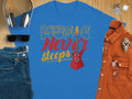 Garment Graphics blue 100% cotton t-shirt showcases a red electric guitar and the phrase Music Never Sleeps, capturing Graphic T-Shirts essence. Its styled in a flat lay with headphones, sunglasses, jeans, a brown shirt, wristwatch, and keys on a wooden surface.