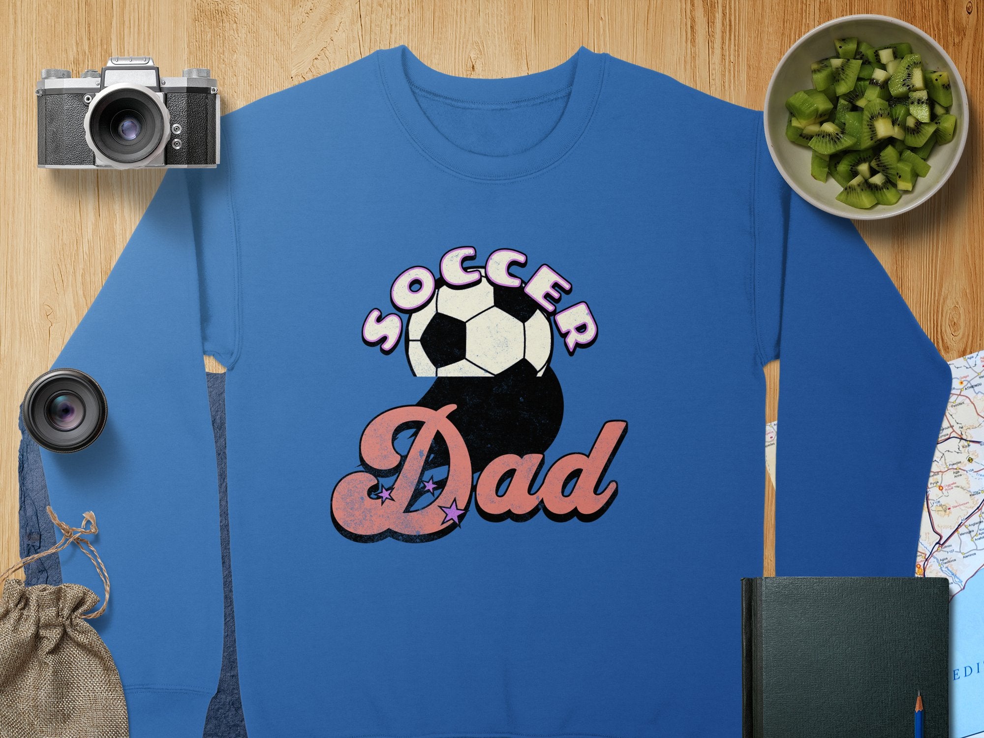 Laid on a wooden surface, the Garment Graphics blue Soccer Dad graphic print sweatshirt features a retro design and soccer ball, surrounded by a camera, lens, green fruit in a bowl, burlap bag, closed book, and map.