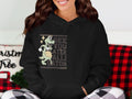 A woman wears a Garment Graphics classic-fit black hoodie featuring a gator and See You Later Alligator text. She smiles, her hair down, amidst festive Christmas decorations and red-and-black checkered fabric.