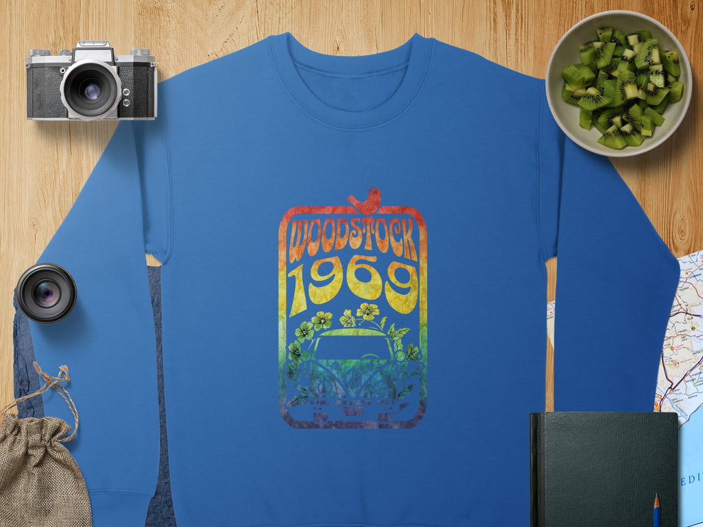 A blue Garment Graphics sweatshirt features a vibrant Woodstock 1969 design, capturing retro vibes with flowers, a car, and a bird. It sits on a wooden surface alongside props like a camera, lens, burlap bag, kiwi bowl, notebook, and map.
