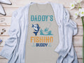 A gray Graphic Print T-Shirt by Garment Graphics, featuring Daddys Fishing Buddy and an image of fishing from a boat with a large fish, is styled with a light blue cardigan on wood decorated with white flowers and jewelry, making it the perfect gift for fishing enthusiasts.