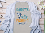 A gray Graphic Print T-Shirt by Garment Graphics, featuring Daddys Fishing Buddy and an image of fishing from a boat with a large fish, is styled with a light blue cardigan on wood decorated with white flowers and jewelry, making it the perfect gift for fishing enthusiasts.