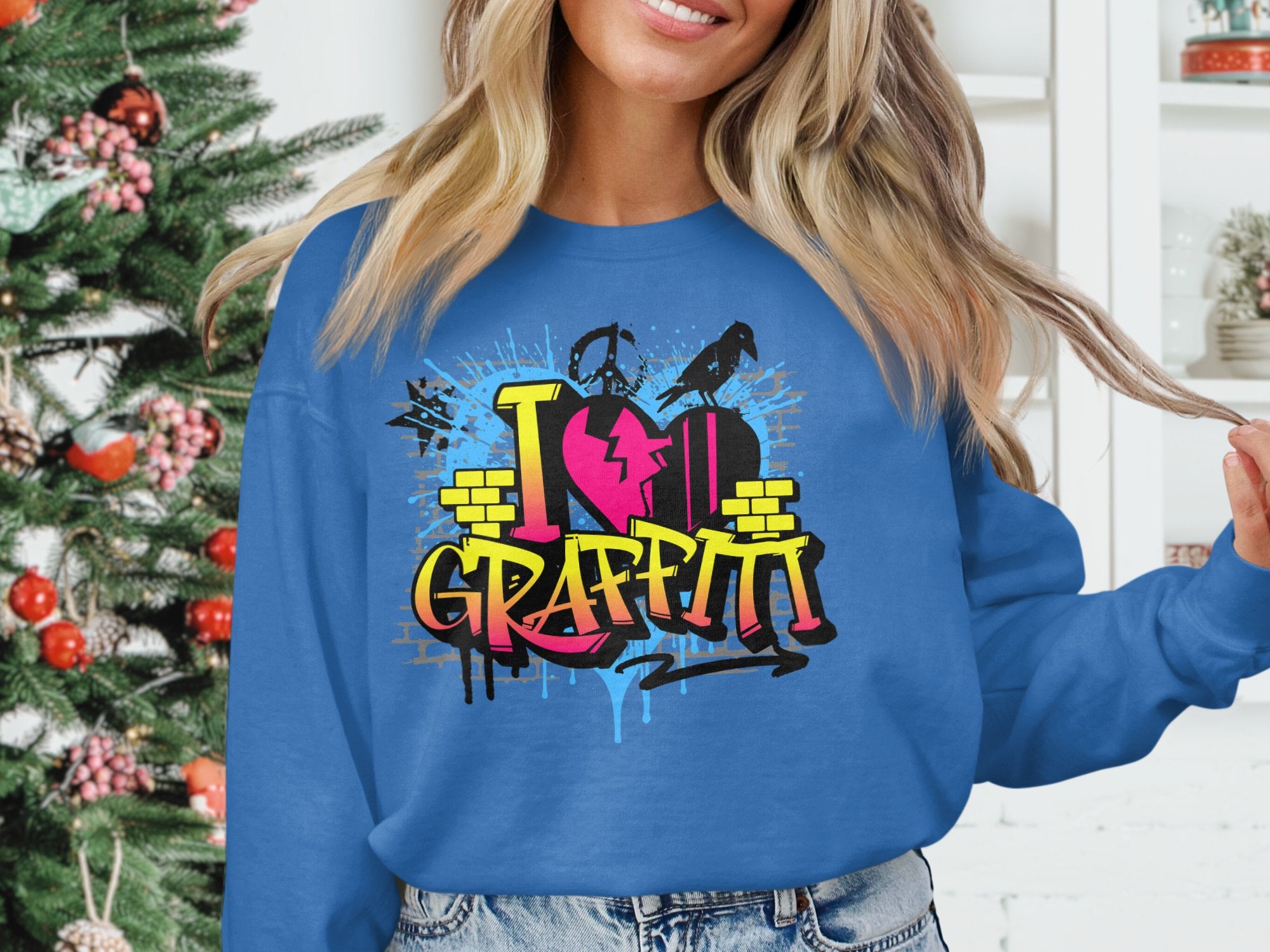 A person in a Garment Graphics sweatshirt with I Heart Graffiti in vibrant lettering stands by a decorated Christmas tree, perfectly complementing the urban style of the colorful shelves behind.