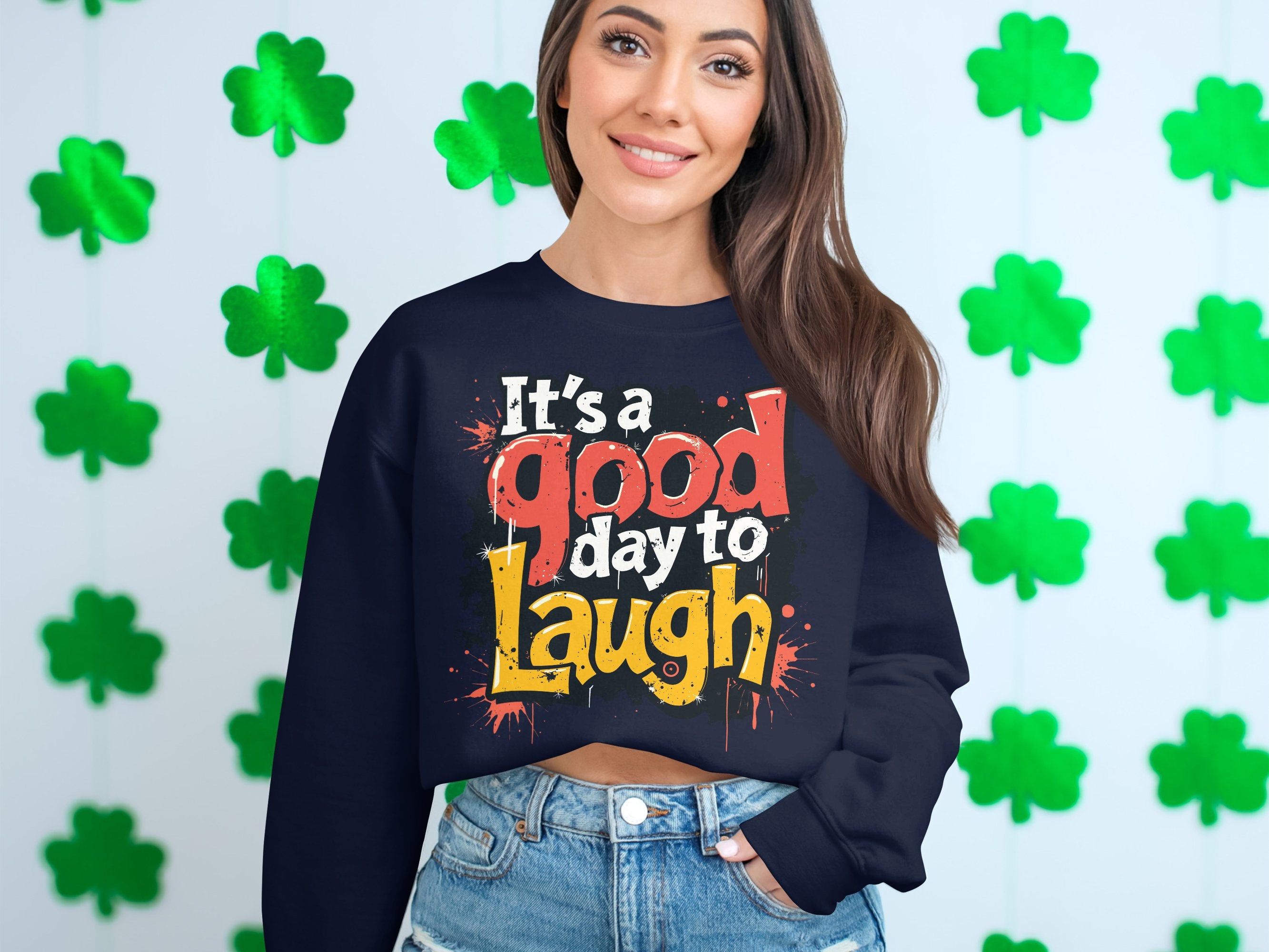 A person with long brown hair smiles, showcasing Garment Graphics dark graphic printed unisex sweatshirt that reads Its a good day to Laugh in colorful letters. They pair it with denim shorts, and green shamrocks decorate the background, enhancing the playful feel.