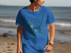 Someone wearing a Royal Mail - 24 Tracked classic fit blue graphic t-shirt with Lucky Just Blessed Shamrock printed on it stands on the beach, with the ocean and sand extending behind them.