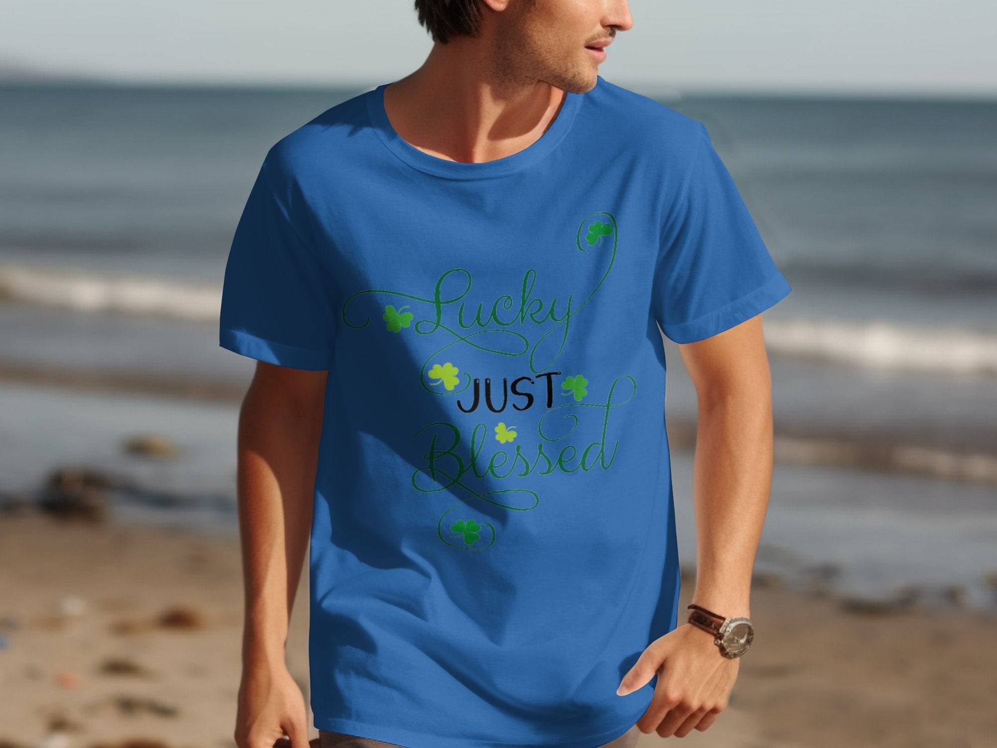 Someone wearing a Royal Mail - 24 Tracked classic fit blue graphic t-shirt with Lucky Just Blessed Shamrock printed on it stands on the beach, with the ocean and sand extending behind them.