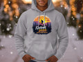 A person sports a Garment Graphics gray hiking hoodie with a sunset mountain and hiker silhouette design. The text reads, Retired 2025. Not My Problem Anymore. Snowfall sets the scene in the background.