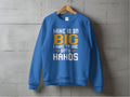 A bold blue sweatshirt by Garment Graphics hangs on a wooden hanger against a gray concrete wall, featuring the eye-catching white and yellow text: MINE IS SO BIG I HAVE TO USE BOTH HANDS.