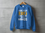 A bold blue sweatshirt by Garment Graphics hangs on a wooden hanger against a gray concrete wall, featuring the eye-catching white and yellow text: MINE IS SO BIG I HAVE TO USE BOTH HANDS.