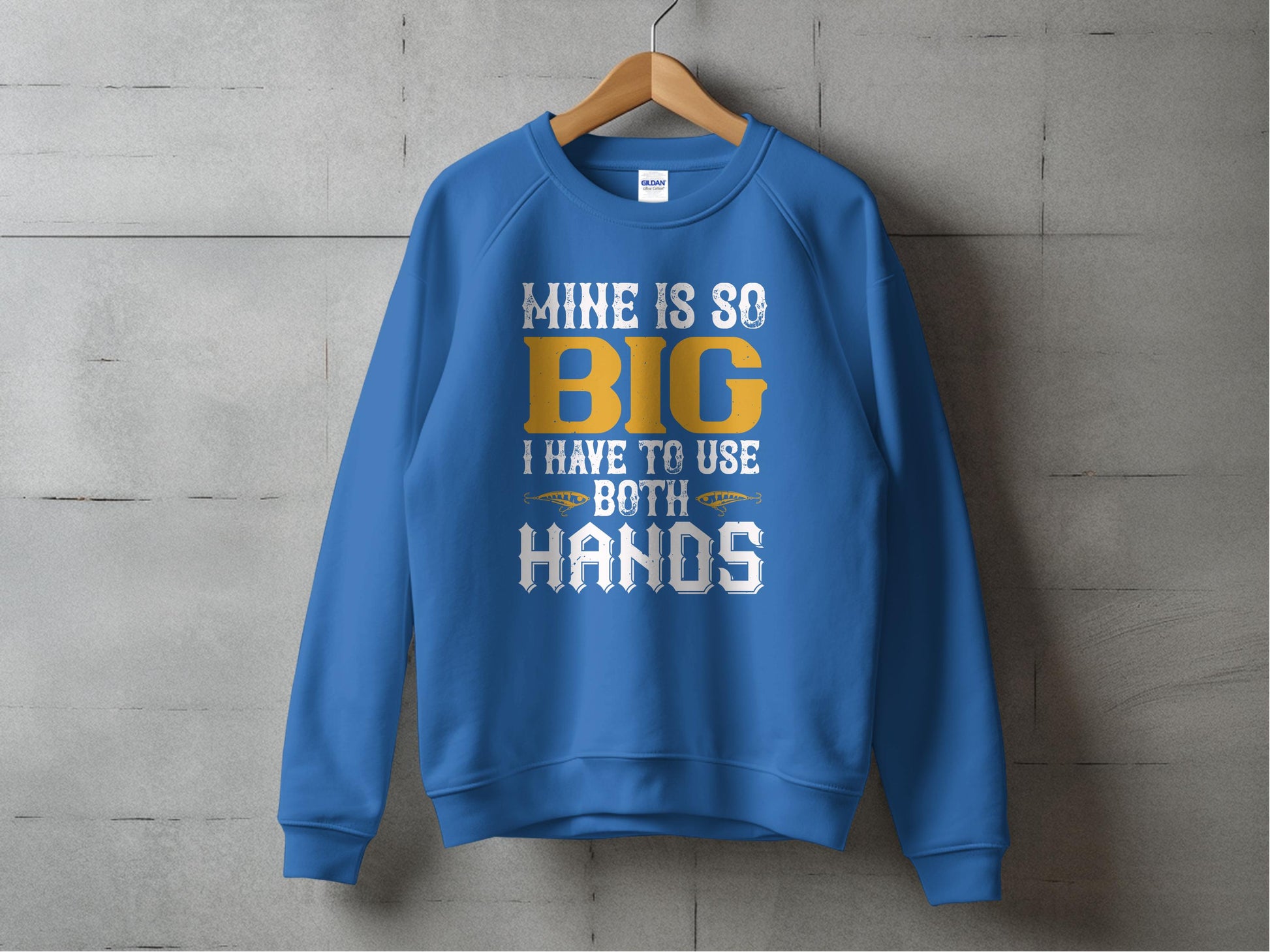 A bold blue sweatshirt by Garment Graphics hangs on a wooden hanger against a gray concrete wall, featuring the eye-catching white and yellow text: MINE IS SO BIG I HAVE TO USE BOTH HANDS.
