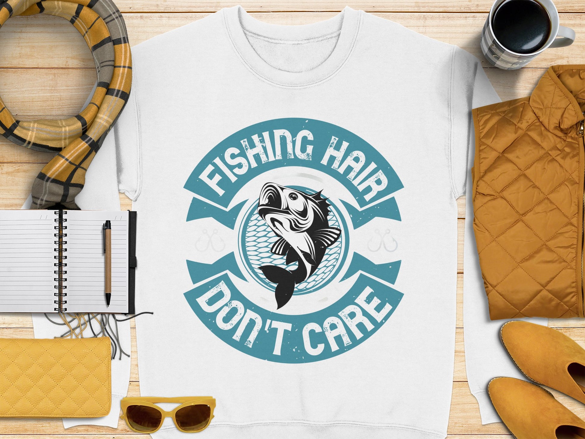 Garment Graphics offers a white T-shirt with Fishing Hair Dont Care text and a fish graphic, ideal for fishing enthusiasts. Displayed on wood with a notebook, pen, yellow sunglasses, purse, plaid scarf, black coffee cup, and mustard jacket in the background.