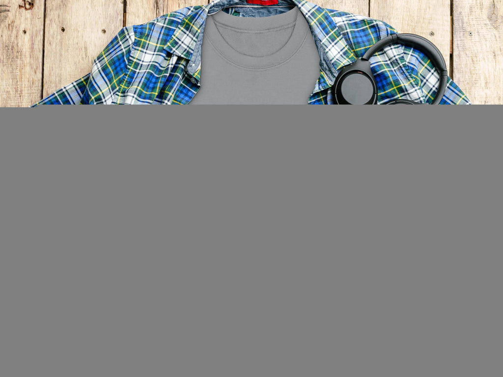A Garment Graphics gray T-shirt with a Happy Patricks Day print of a frothy beer mug and shamrocks is set against a blue and green plaid shirt. Its surrounded by a camera, headphones, and coffee on wood, ideal for stylish St. Patricks Day celebrations.
