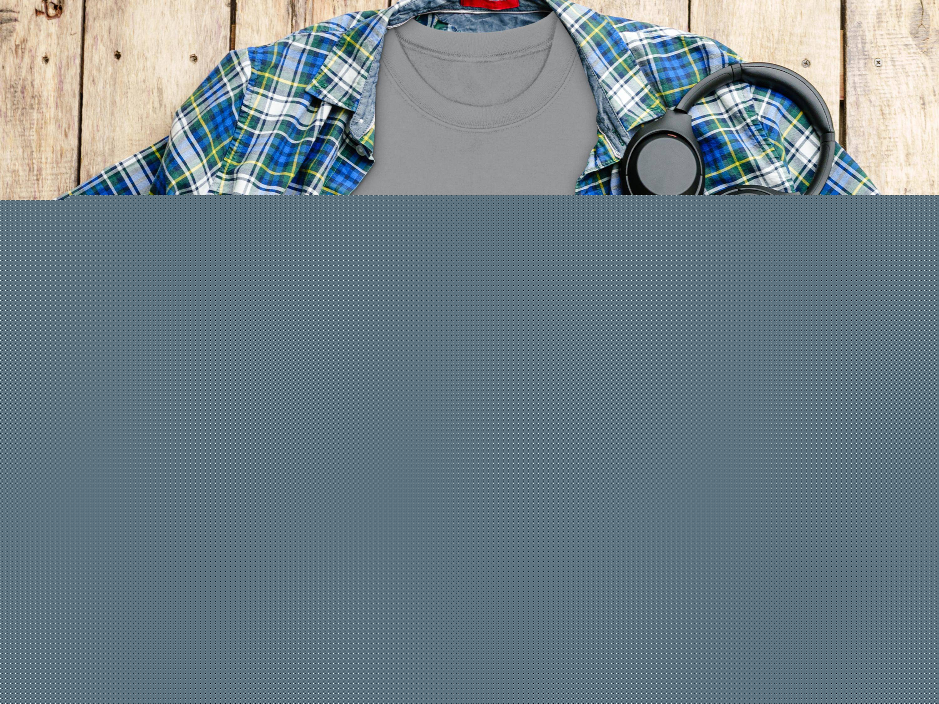 A Garment Graphics gray T-shirt with a Happy Patricks Day print of a frothy beer mug and shamrocks is set against a blue and green plaid shirt. Its surrounded by a camera, headphones, and coffee on wood, ideal for stylish St. Patricks Day celebrations.