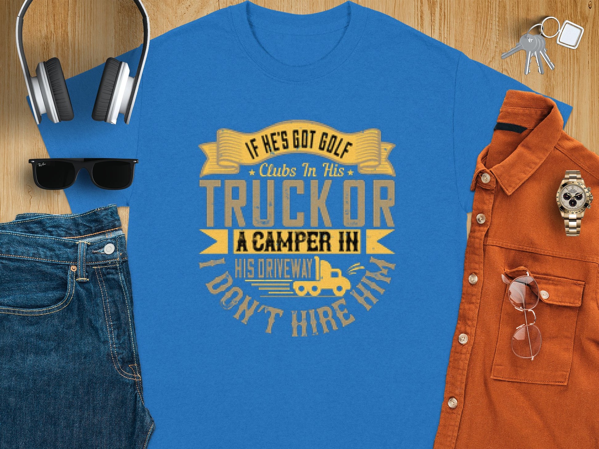 A blue T-shirt from Garment Graphics humorously declares, If hes got golf clubs in his truck or a camper in his driveway, I dont hire him. Nearby on the wooden surface are headphones, sunglasses, a brown shirt, watch, and blue jeans—ideal for fans of golfing and camping humor.