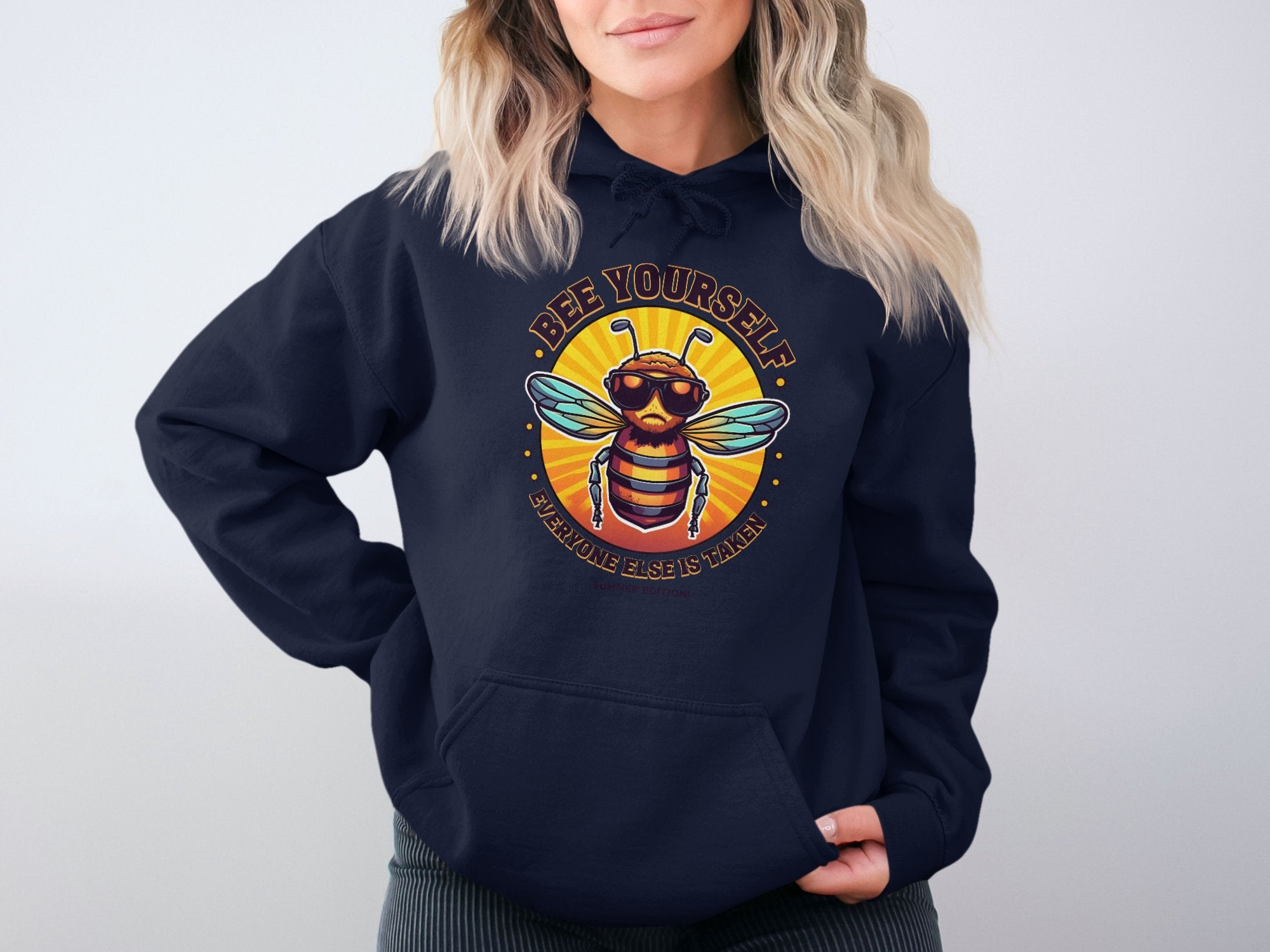 A person models the Bee Yourself hoodie by Garment Graphics, featuring a bee with sunglasses and the text Bee Yourself above and Everyone Else is Taken below. With wavy blonde hair against a plain backdrop, this hoodie makes a striking impression.