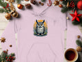 The Garment Graphics Fierce Owl Illustration Hoodie features a medium-heavy fabric in light pink with an owl graphic on an orange backdrop, set amidst Christmas decor like pine branches, ornaments, pinecones, and a steaming coffee mug, offering unique style on any wooden surface.